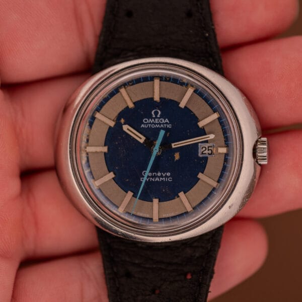 A hand holding an Omega Automatic Geneve Dynamic Ref. 166.107 Cal. 1421 wristwatch with a blue and silver dial.
