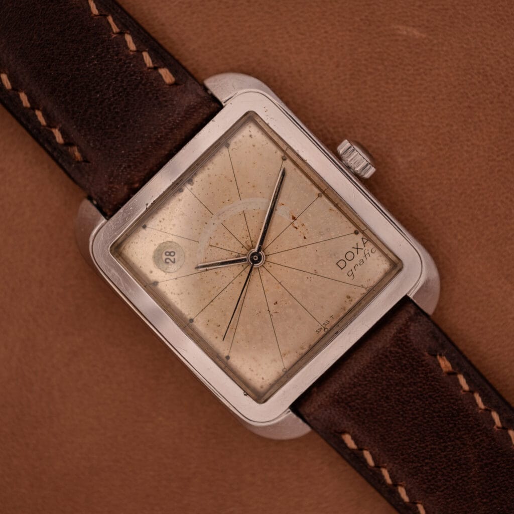 A Doxa Graphic Ref. 11728-4 with a stainless steel square case and brown leather strap rests on a brown surface, highlighting the watch's timeless elegance.