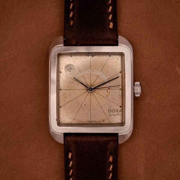 A Doxa Graphic Ref. 11728-4 watch featuring a stainless steel square case and brown leather strap, shown on a tan background.