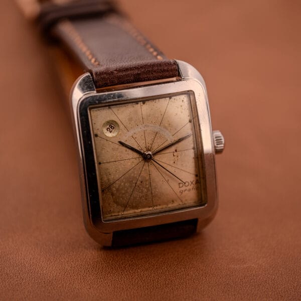 A Doxa Graphic Ref. 11728-4 wristwatch with a stainless steel square case, worn dial, and brown leather strap rests on a brown surface.