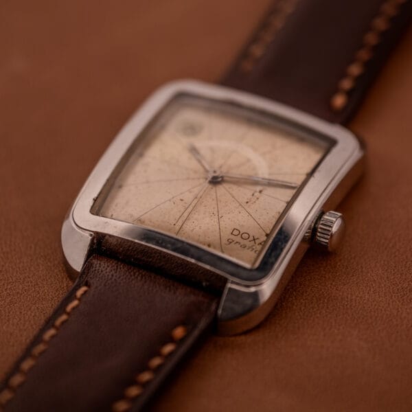 The Doxa Graphic Ref. 11728-4, featuring a stainless steel square case and brown leather strap, rests elegantly on a brown surface.