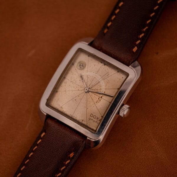 The Doxa Graphic Ref. 11728-4 wristwatch features a stainless steel square case and a brown leather strap, displayed on a brown surface showing the time 3:48.