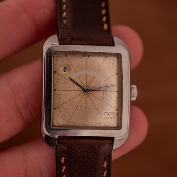 The Doxa Graphic Ref. 11728-4 features a stainless steel square case, a weathered patina dial, and a brown leather strap for a classic vintage look.