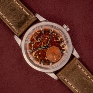An Omega Automatic 2491 "Pre-Seamaster" Tropical Dial Cal. 28.10 RA with a tan leather strap sits on a burgundy surface, radiating vintage charm.