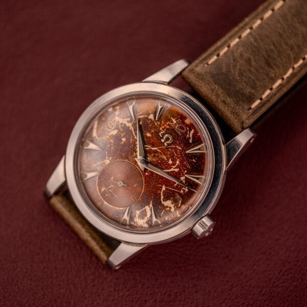 An Omega Automatic 2491 "Pre-Seamaster" Tropical Dial Cal. 28.10 RA wristwatch with a brown leather strap and weathered tropical dial rests on a burgundy surface.