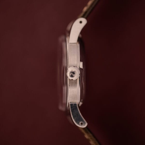 Side view of a silver Omega Automatic 2491 "Pre-Seamaster" Tropical Dial Cal. 28.10 RA wristwatch with crown, set against a dark red background.