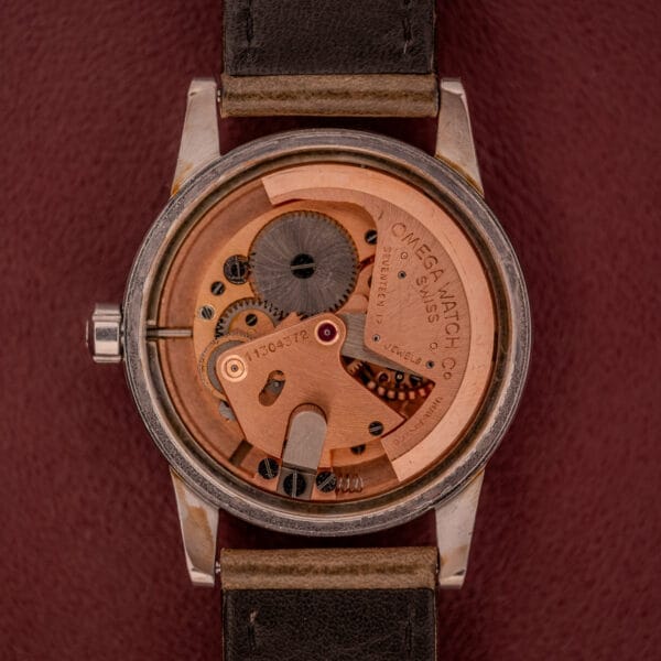 The Omega Automatic 2491 "Pre-Seamaster" Tropical Dial Cal. 28.10 RA watch with exposed movement and black strap is displayed on a textured maroon background.