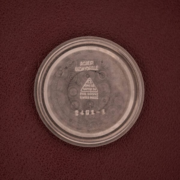 A round metal Omega Automatic 2491 "Pre-Seamaster" Tropical Dial Cal. 28.10 RA watch case back rests on a textured brown surface, displaying engraved text and numbers—an intriguing vintage collector’s piece.