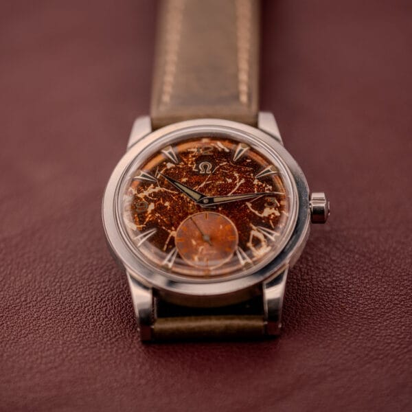 The Omega Automatic 2491 "Pre-Seamaster" features a brown tropical dial and leather strap, showcased on a burgundy surface.