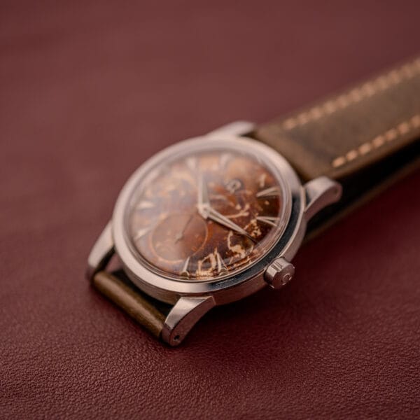 An Omega Automatic 2491 "Pre-Seamaster" Tropical Dial Cal. 28.10 RA with a leather strap rests on a reddish-brown surface, showcasing timeless vintage style.
