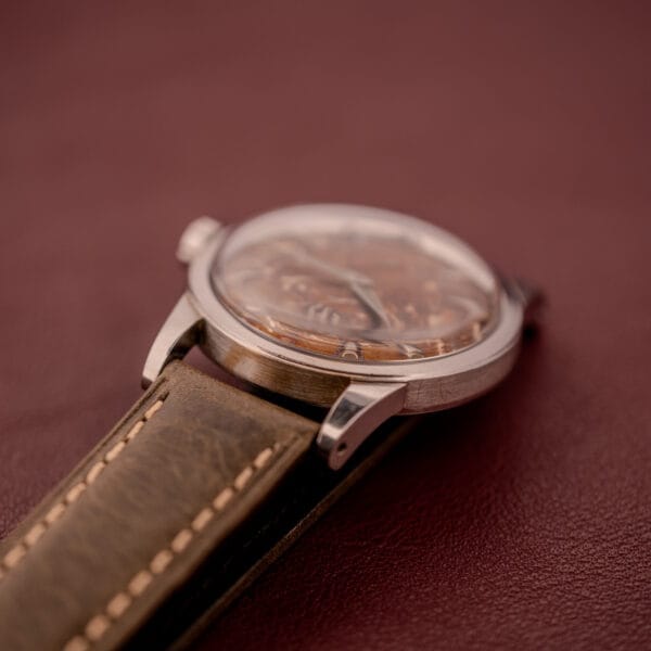 Close-up of an Omega Automatic 2491 "Pre-Seamaster" Tropical Dial Cal. 28.10 RA with a brown leather strap, shown on a maroon surface.