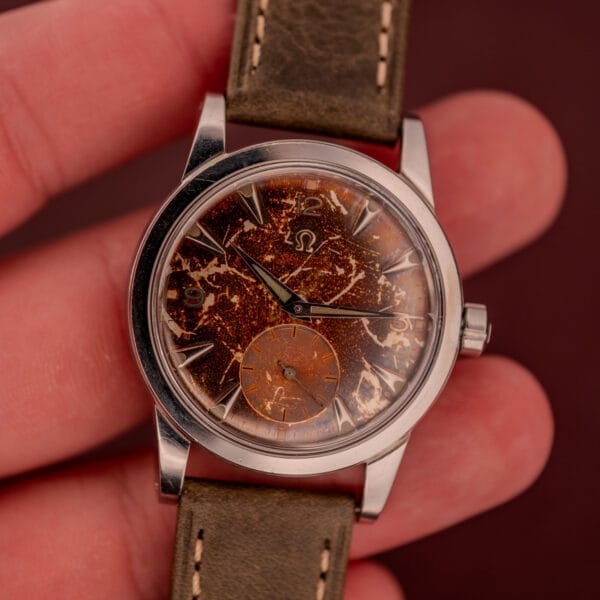 A hand holds an Omega Automatic 2491 "Pre-Seamaster" Tropical Dial Cal. 28.10 RA wristwatch with a distressed tropical dial and olive leather strap.