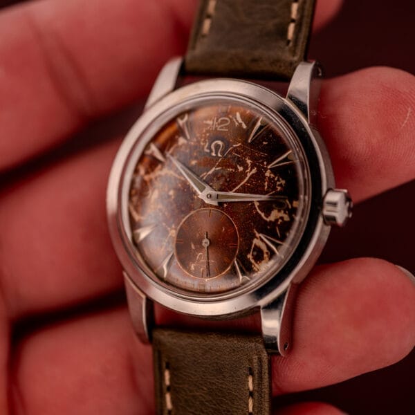 A hand holding an Omega Automatic 2491 "Pre-Seamaster" Tropical Dial Cal. 28.10 RA, featuring a worn brown face and paired with a green leather strap.