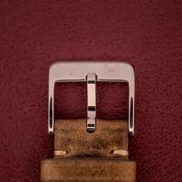 A close-up of a silver buckle on a brown leather strap, set against maroon—an elegant detail from the Omega Automatic 2491 "Pre-Seamaster" Tropical Dial Cal. 28.10 RA.