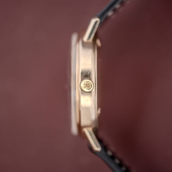 Side view of the Girard Perregaux Quartz Date 3696 Champagne Dial Cal. ETA 940 wristwatch with crown logo, shown against a blurred maroon background.