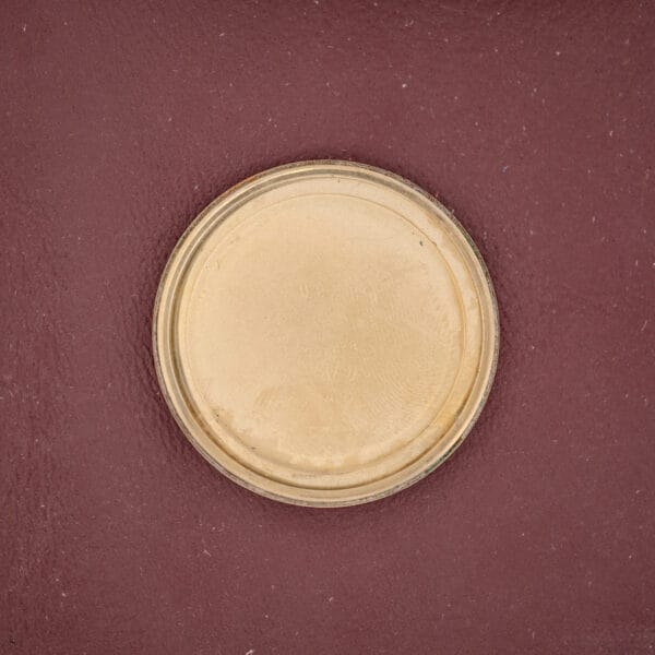 A round, gold metal lid inspired by the Girard Perregaux Quartz Date 3696 Champagne Dial rests on a textured maroon surface.