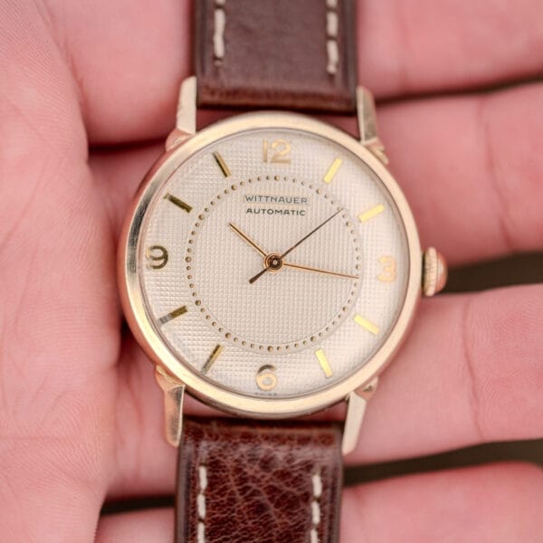 A close-up of a Wittnauer Automatic Waffle Dial Ref. 2023 wristwatch with a 10k GF case and brown leather strap, featuring Cal. 11ARB, held in a person's hand.