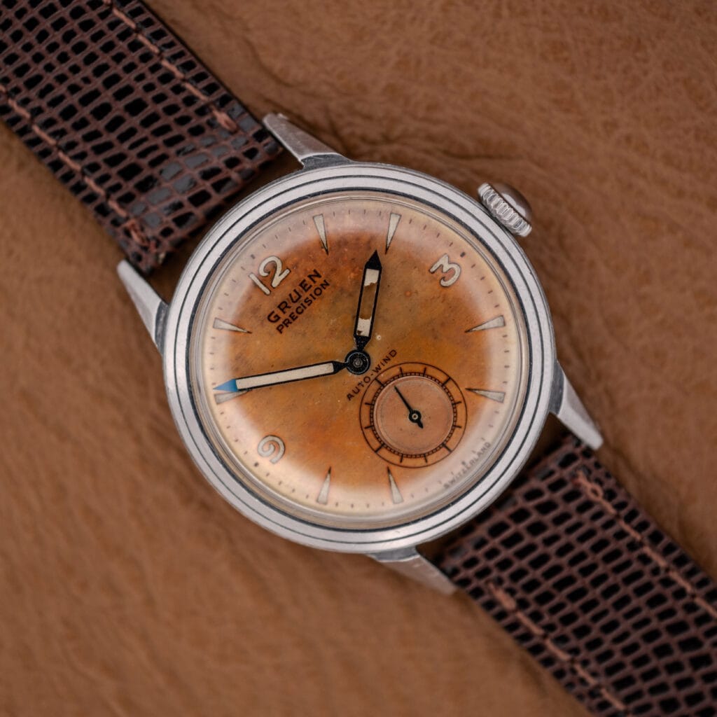 A Gruen Precision Auto-Wind Copper Dial in Stainless Steel Cal. 400 wristwatch with a brown leather strap rests on a tan textured surface.