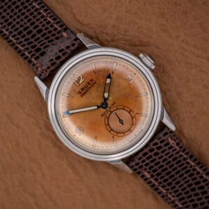 A Gruen Precision Auto-Wind Copper Dial in Stainless Steel Cal. 400 wristwatch with a brown leather strap rests on a tan textured surface.