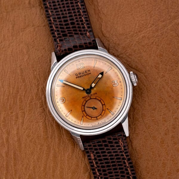 The Gruen Precision Auto-Wind Copper Dial in Stainless Steel Cal. 400 wristwatch, with a brown leather strap, is elegantly displayed on a textured brown surface.