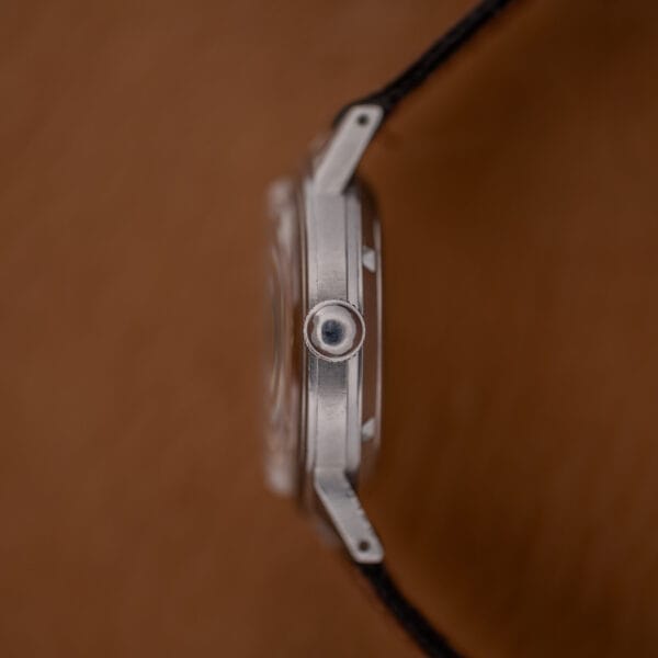 Side view of the Gruen Precision Auto-Wind Copper Dial in Stainless Steel Cal. 400, featuring a silver crown, black strap, and copper dial against a blurred brown background.