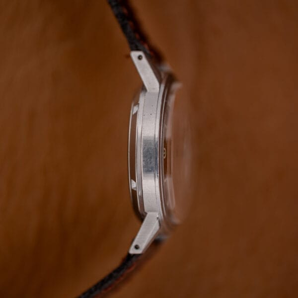 Side view of the Gruen Precision Auto-Wind Copper Dial in Stainless Steel Cal. 400 wristwatch with a black strap, shown against a brown background.