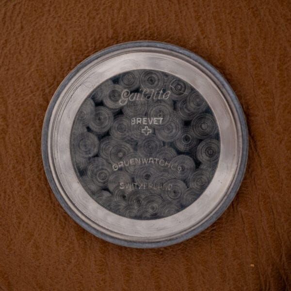 The round stainless steel case back with engraved text rests on a brown textured surface, highlighting the quality craftsmanship of the Gruen Precision Auto-Wind Copper Dial in Stainless Steel Cal. 400.