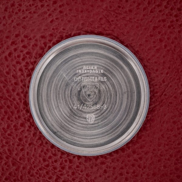 The Tissot Seastar Ref. 42568 showcases a round stainless steel case back with engraved text, set against a textured red background. Features include a matte black dial and reliable Cal. 781-1 movement for lasting quality.