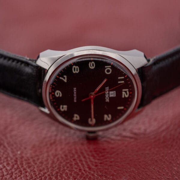 A Tissot Seastar Ref. 42568 wristwatch with a matte black dial and stainless steel case rests on a textured red surface.