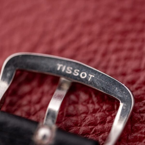 A close-up of the Tissot Seastar Ref. 42568 buckle on a textured red background, showcasing its refined stainless steel Cal. 781-1 finish and matte black dial.