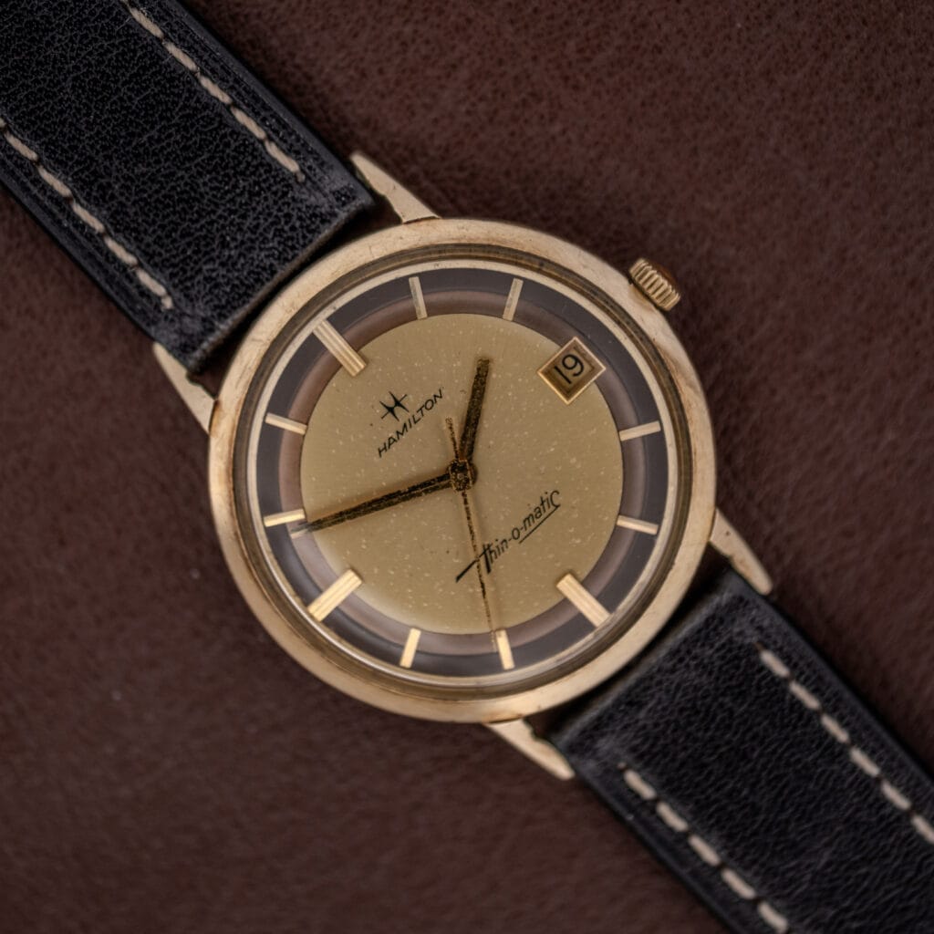 Hamilton Thin-O-Matic Brown & Gold Dial Automatic Cal. 624 wristwatch with black leather strap, displayed on a brown background.