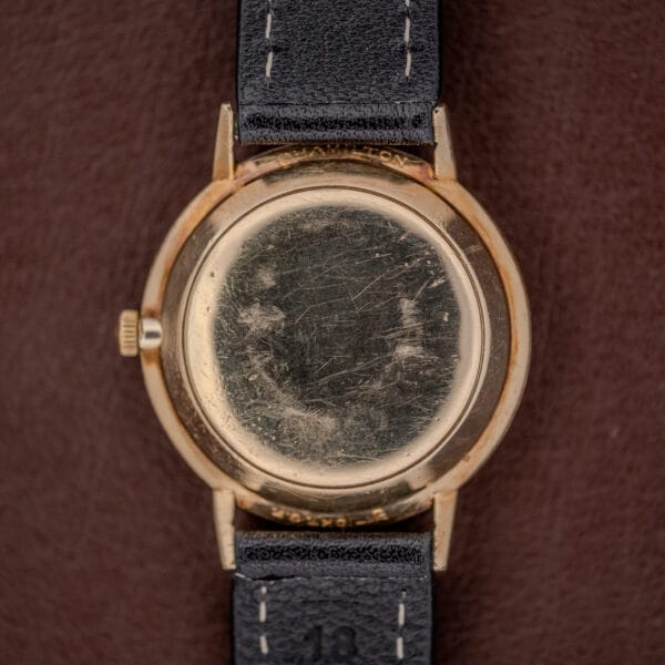 A Hamilton Thin-O-Matic Brown & Gold Dial Automatic Cal. 624 watch with a scratched gold case and black leather strap, displayed from the back on a brown surface.