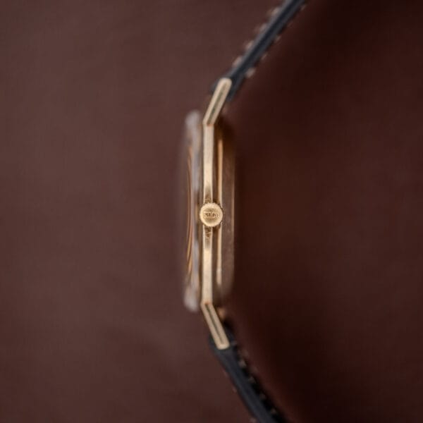 A close-up side view of the Hamilton Thin-O-Matic Brown & Gold Dial Automatic Cal. 624 gold wristwatch with a black strap, highlighting its crown and stylish brown & gold dial against a brown background.