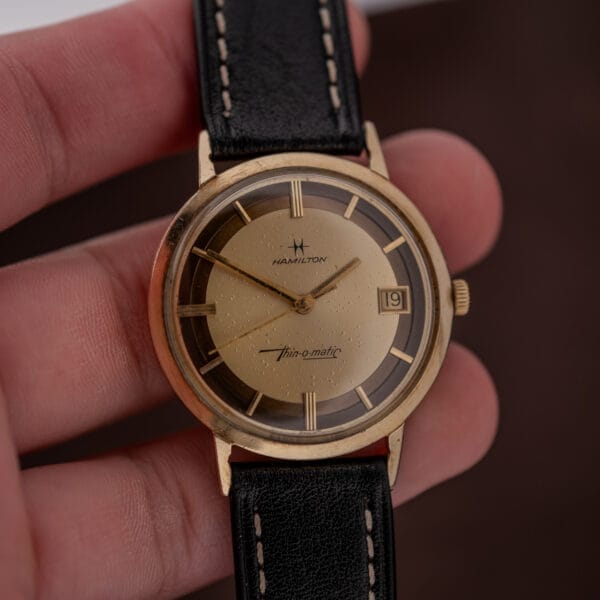 A hand holds the Hamilton Thin-O-Matic Brown & Gold Dial Automatic Cal. 624, featuring a brown and gold dial, black leather strap, and date window at 3 o’clock.
