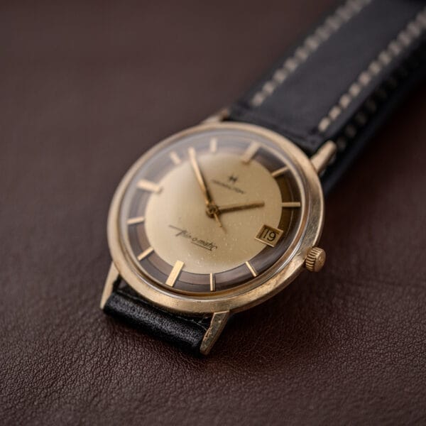 A Hamilton Thin-O-Matic Brown & Gold Dial Automatic Cal. 624 wristwatch with a black leather strap rests on a brown surface.