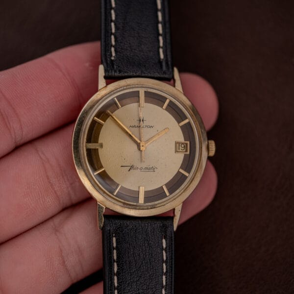 A hand holds a Hamilton Thin-O-Matic Brown & Gold Dial Automatic Cal. 624 watch with a black leather strap and date display.