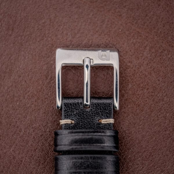 Close-up of a silver belt buckle on a black leather belt, set against a brown textured background inspired by the refined elegance of the Hamilton Thin-O-Matic Brown & Gold Dial Automatic Cal. 624.