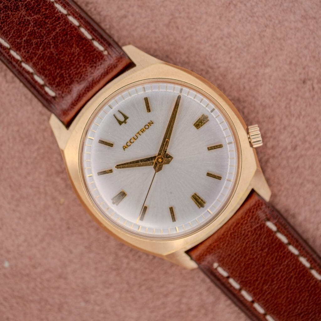 A Bulova Accutron Gold Cushion Case with Silver Sunburst Dial wristwatch featuring a brown leather strap rests on a pink textured surface.
