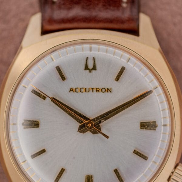 Close-up of a Bulova Accutron Gold Cushion Case wristwatch, featuring a silver sunburst dial and brown leather strap.