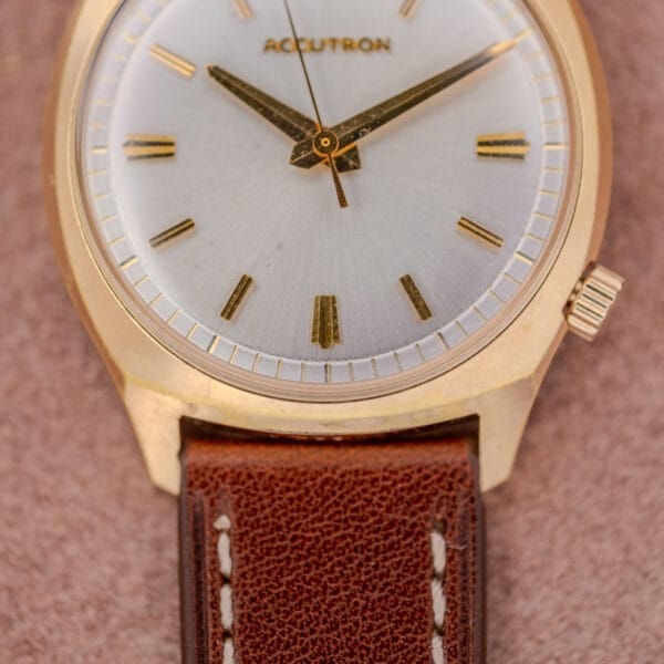 Bulova Accutron Gold Cushion Case with Silver Sunburst Dial and brown leather strap rests on a soft tan surface, displaying the time 8:20.