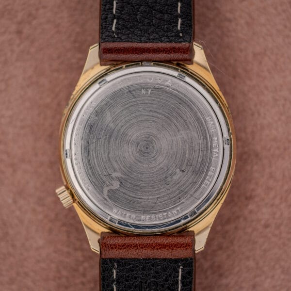Back of a worn Bulova Accutron Gold Cushion Case with Silver Sunburst Dial wristwatch featuring a black leather strap and circular scratches on the metal case.