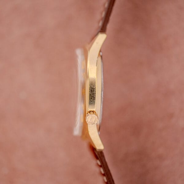 Side view of a Bulova Accutron Gold Cushion Case with Silver Sunburst Dial and brown leather strap on a soft pink background.