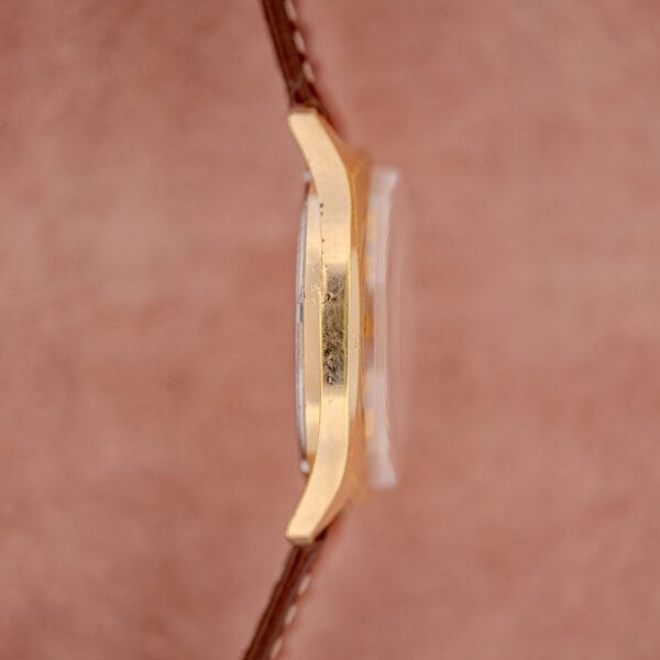 Side view of the Bulova Accutron Gold Cushion Case with Silver Sunburst Dial, featuring a brown leather strap, displayed against a soft pink background.