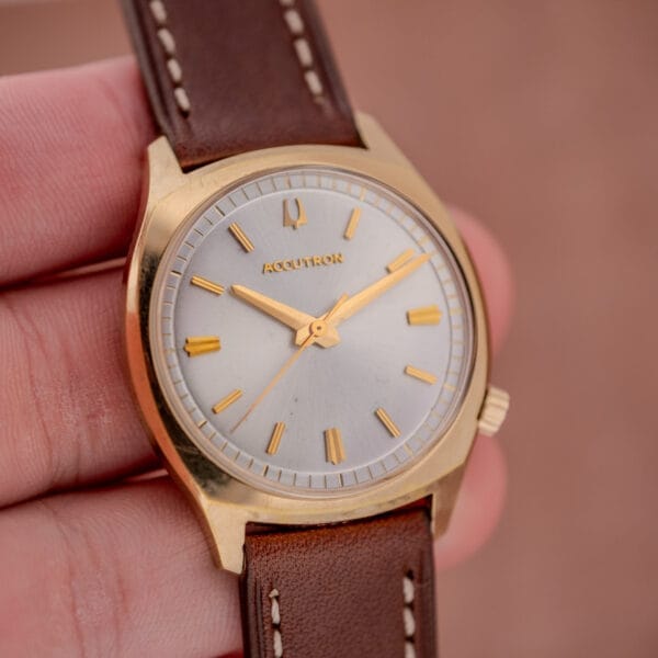 A hand holding the Bulova Accutron Gold Cushion Case with a striking silver sunburst dial and a brown leather strap.