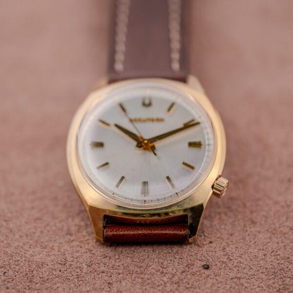 Bulova Accutron Gold Cushion Case with Silver Sunburst Dial wristwatch featuring a brown leather strap, shown on a soft brown surface with the time set to 2:45.