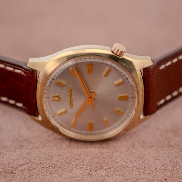 Bulova Accutron Gold Cushion Case with Silver Sunburst Dial wristwatch with a brown leather strap rests on a soft, light brown textured surface.