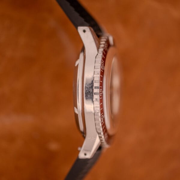 Side view of the silver Wittnauer Professional Chronograph Ref. 7004 Landeron 248 with red bezel and black strap, displayed against a brown background.