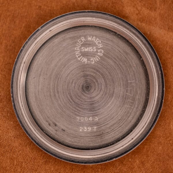 A round metal case back inscribed Wittnauer Watch Co. Inc Swiss and numbers, likely from a Wittnauer Professional Chronograph Ref. 7004 Landeron 248, rests on a brown surface.