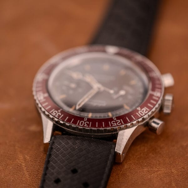 A close-up of a vintage Wittnauer Professional Chronograph Ref. 7004 Landeron 248 with a red bezel and black strap on a brown surface.
