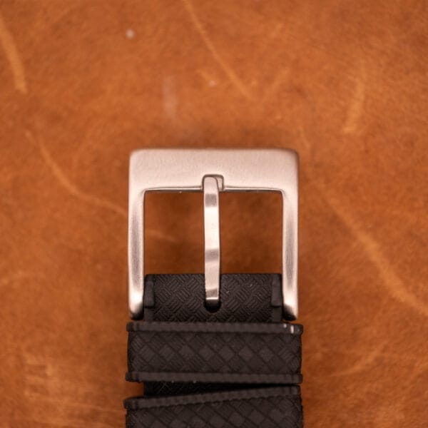 A silver metal belt buckle on a textured black strap lies on brown leather, echoing the sleek style of the Wittnauer Professional Chronograph Ref. 7004 Landeron 248.